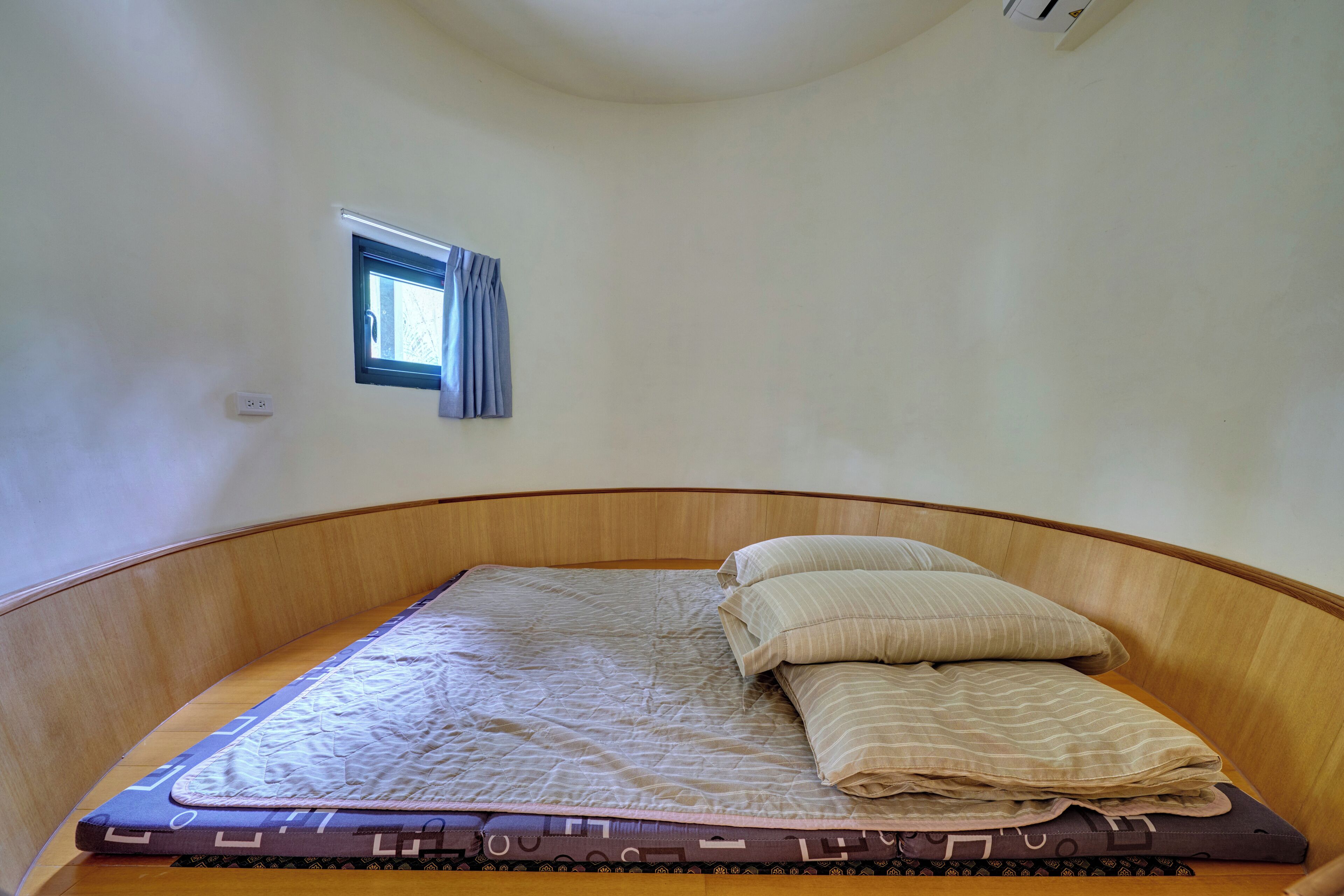 A single bed with a mattress and pillows, a small window with curtains, and a wall-mounted light switch.