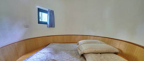 A single bed with a mattress and pillows, a small window with curtains, and a wall-mounted light switch.