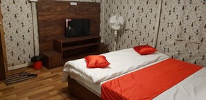 A bedroom with a bed, a TV on a wooden stand, a standing fan, and a red trash bin.