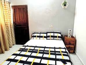 A bedroom with a bed, a wooden door, a small wooden dresser, a potted plant, and a light switch on the wall.