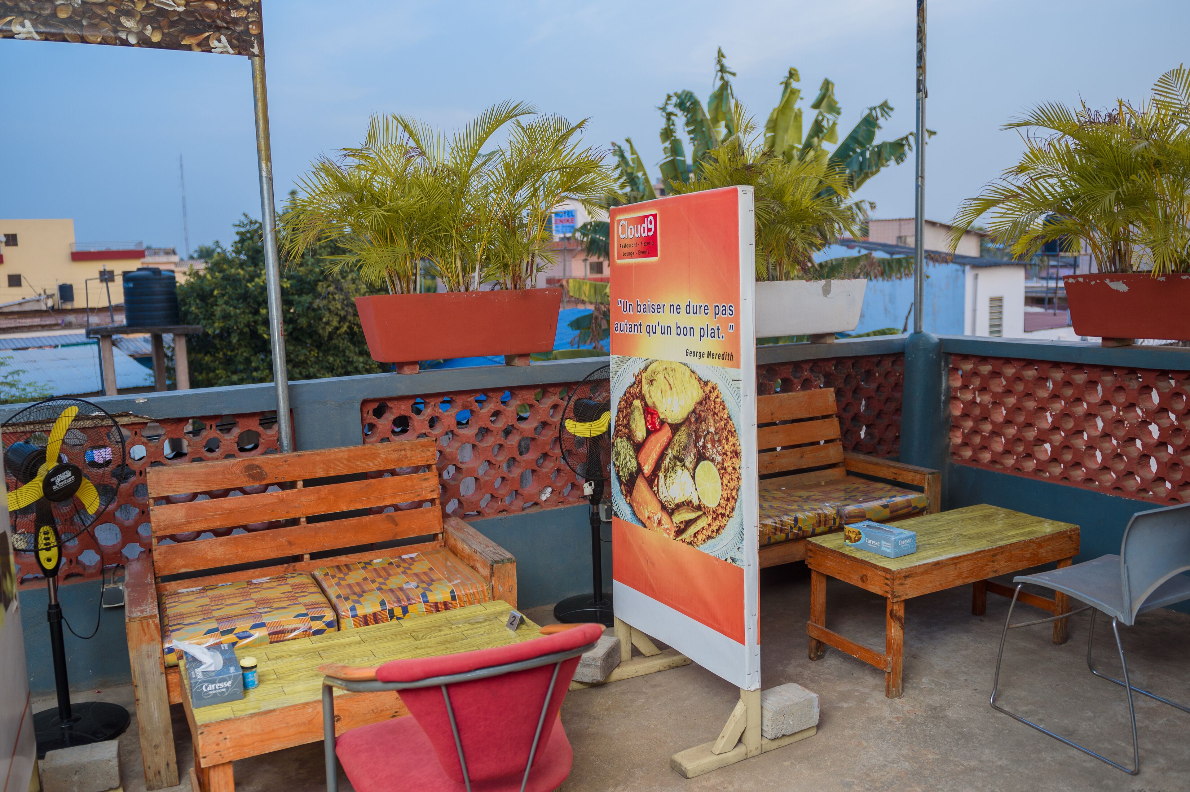 An outdoor seating area with wooden benches and tables, a fan, and a poster.