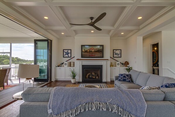 A spacious living room with a fireplace, a ceiling fan, and a large window overlooking a deck.