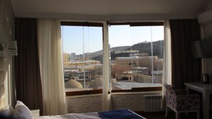 A hotel room with a large window offering a city view, a bed, a chair, and a small table with a kettle. - Old East Boutiques (Baku)