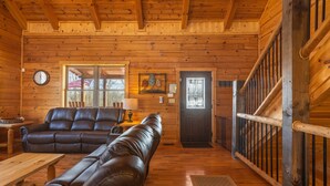 A log cabin interior with a wooden ceiling, leather sofas, a dining table, a staircase, and a door with an American flag.