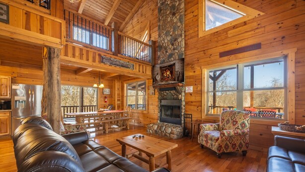 A log cabin interior with a stone fireplace, leather seating, a dining area, and large windows.