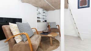Sala de estar con sofá, sillas, una mesa de centro y televisor. Se aprecia una cocina al fondo con armarios y una lámpara colgante.