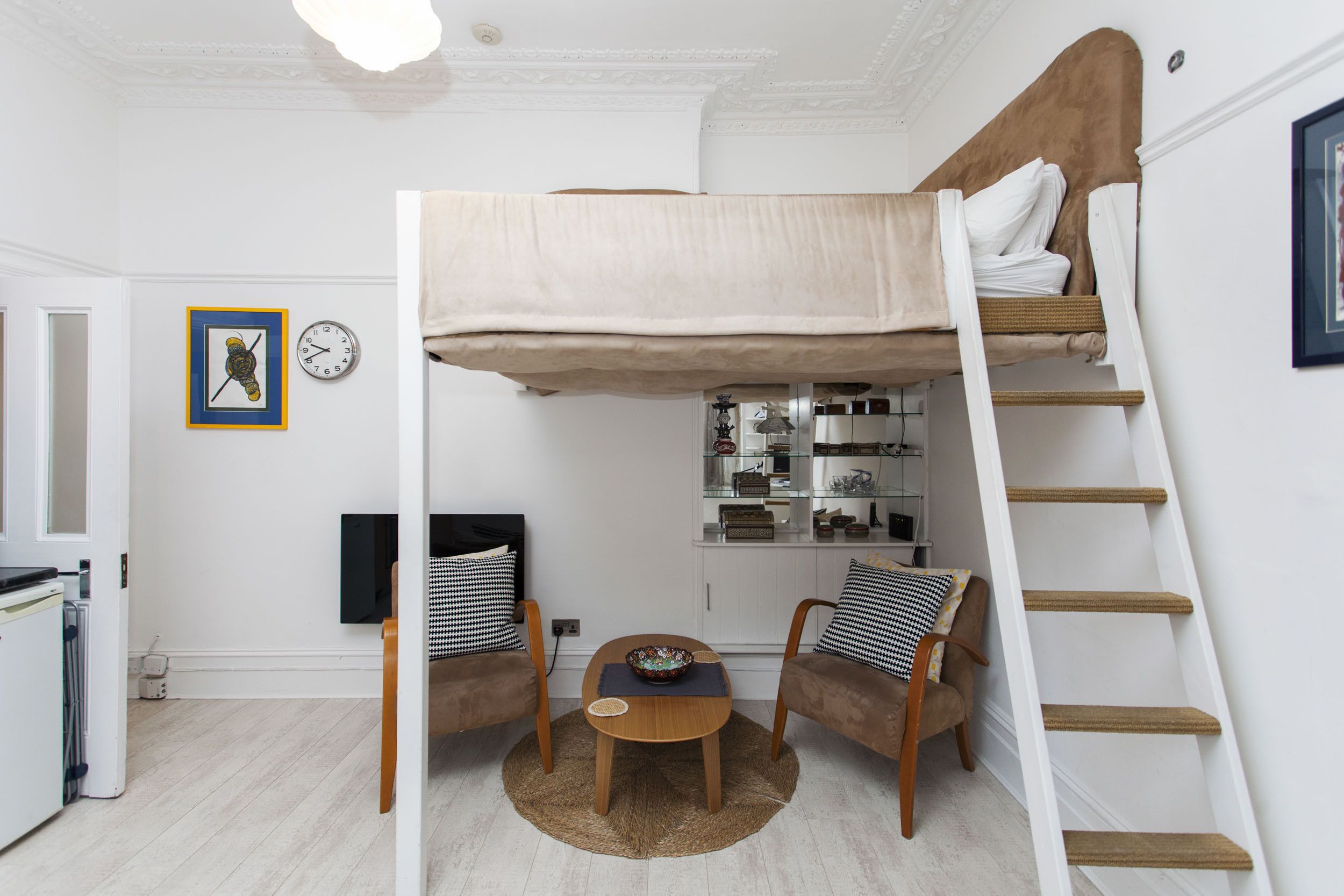A bunk bed with a ladder, a small table with a bowl, two chairs, a clock, and a framed picture on the wall.
