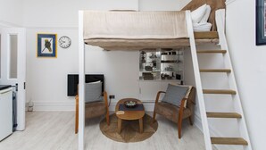 Una cama litera con escalera, una mesita con un cuenco, dos sillas, un reloj y un cuadro en la pared.