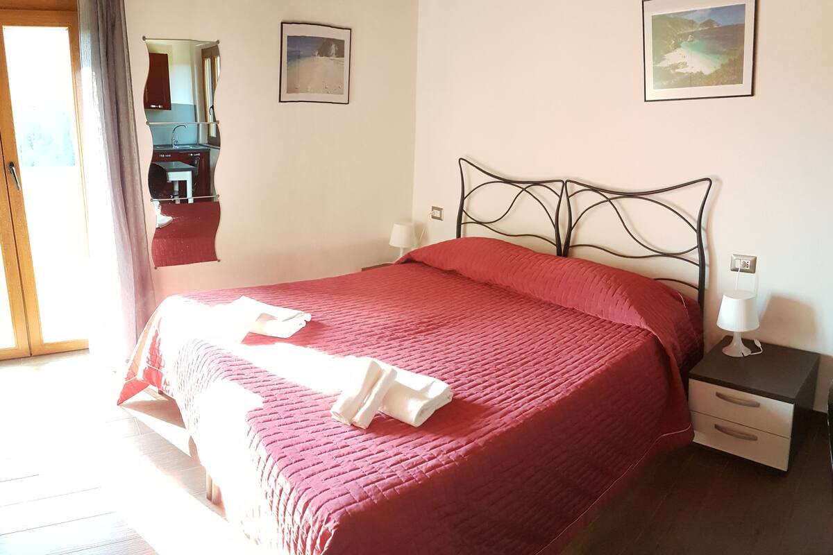 A neatly made bed with a red bedspread and white towels, a bedside table with a lamp, and a small drawer.