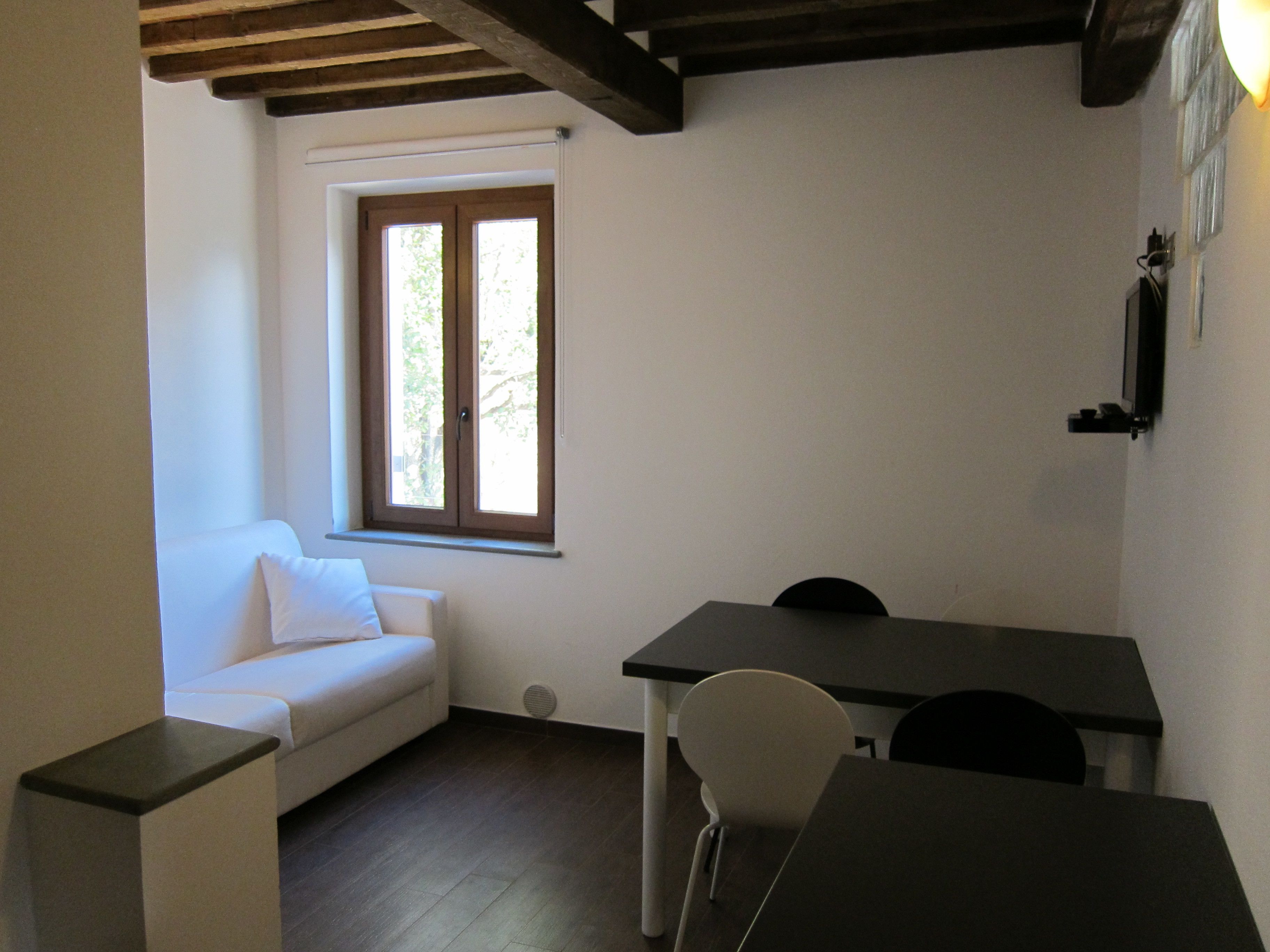 Um quarto pequeno e bem iluminado, com um sofá branco, uma mesa e uma janela com vista para árvores.