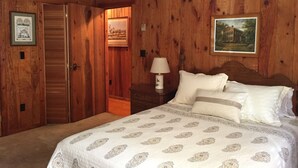 Un dormitorio con paneles de madera, una cama con ropa de cama estampada, una mesita de noche con una lámpara y dos cuadros en las paredes.