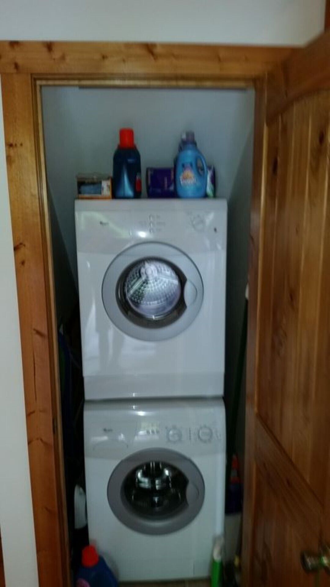 A compact laundry room with a front-loading washer and dryer, wooden cabinets, and various cleaning supplies on top.
