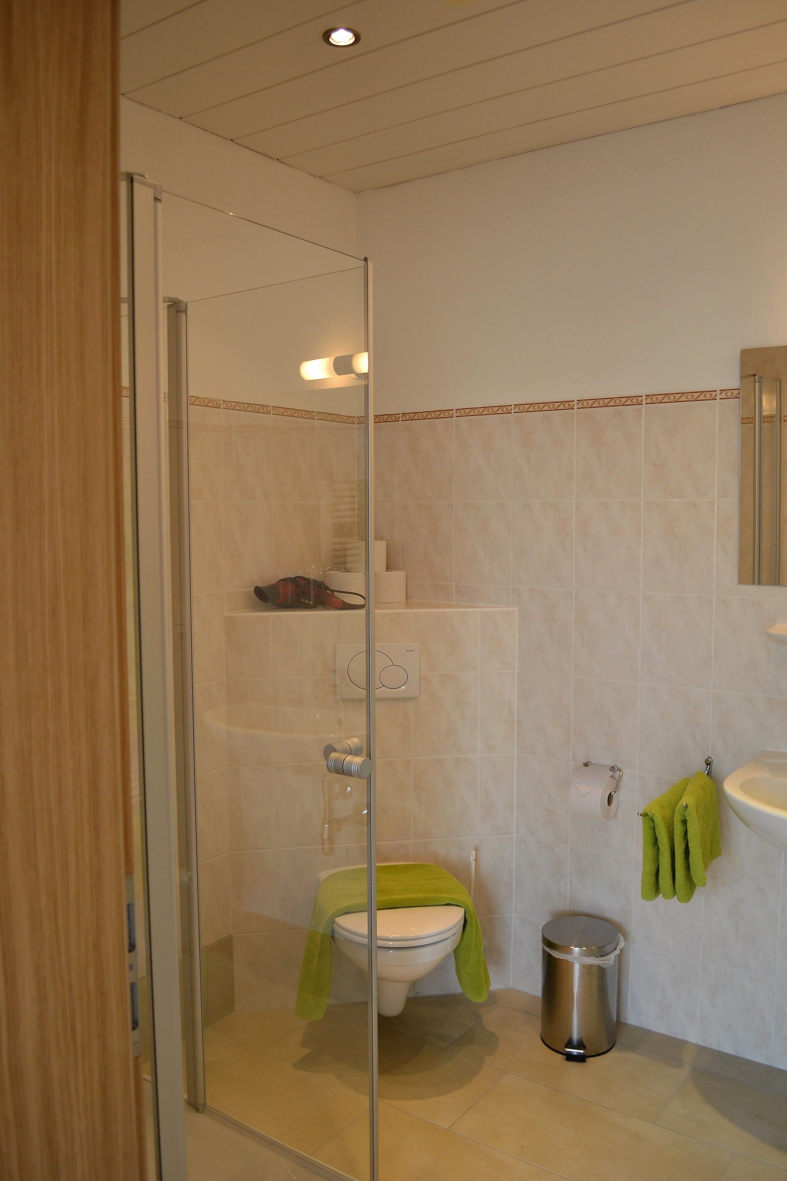 A bathroom with a glass shower enclosure, a toilet, a trash can, and a sink with a green towel.