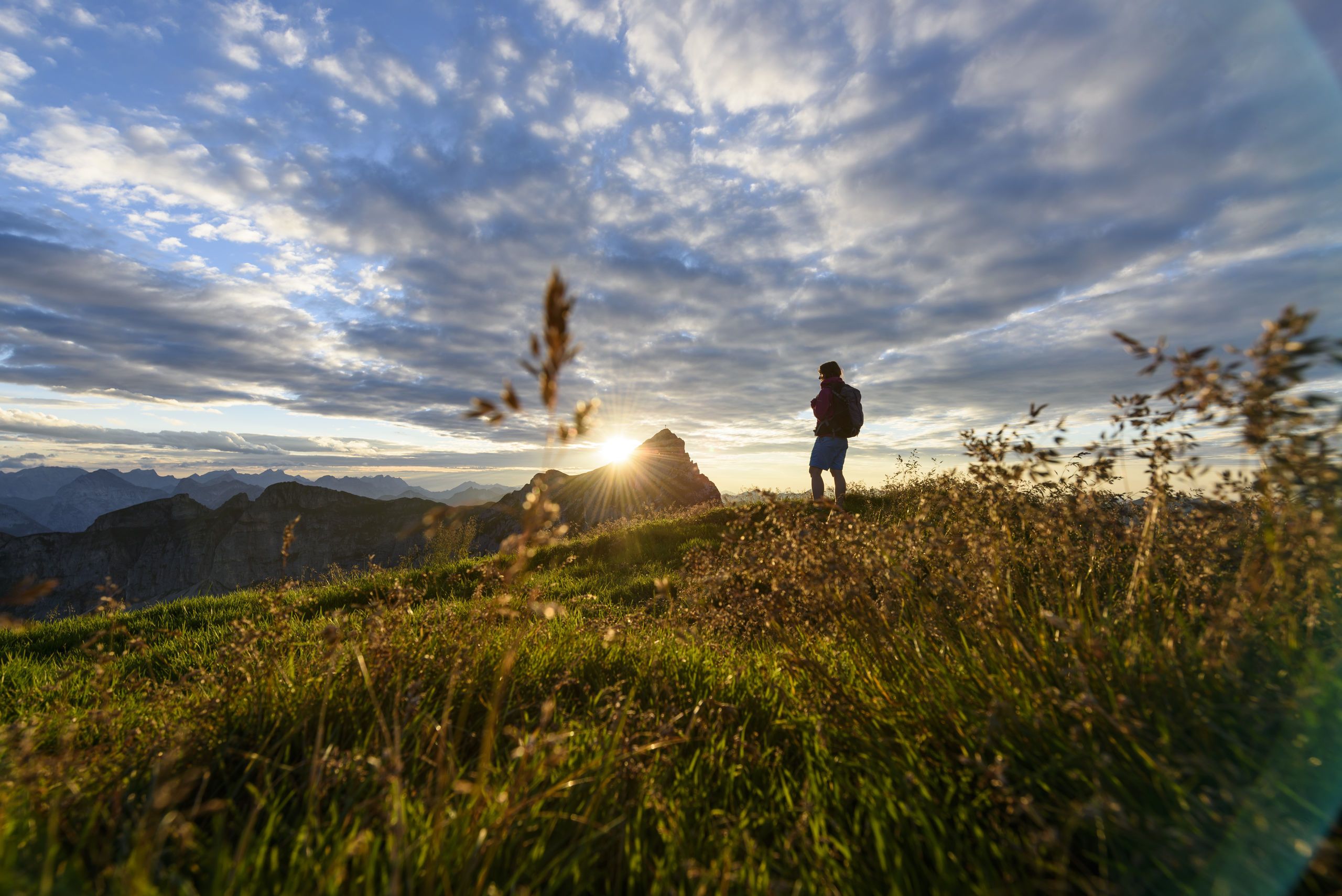 Iemand staat op een grasheuvel, met een bergachtig landschap op de achtergrond en de zon die ondergaat.