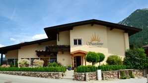 A two-story building with a sign reading "Sunshine Apartments." It features a balcony with flower boxes, a stone wall in front, and a mountain in the background.