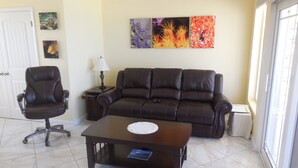 A living room with a brown leather sofa, a black office chair, and a coffee table. There are three framed pictures on the wall and a lamp on a side table. - RazorBrac SeaEsta - Private Beachfront Home (Cayman Brac)