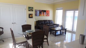 A dining area with a glass table, chairs, and a sofa. There are paintings on the wall, a door, and a window with blinds. - RazorBrac SeaEsta - Private Beachfront Home (Cayman Brac)
