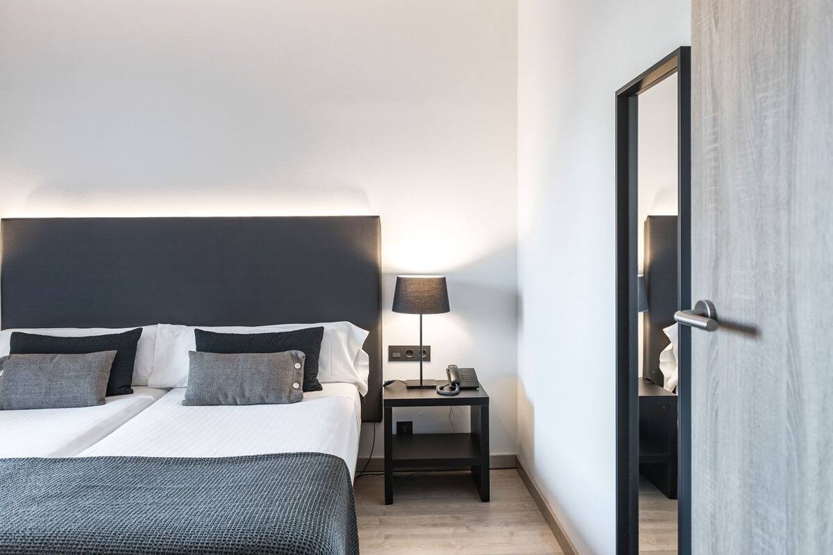 A modern hotel room with a large bed, bedside table with a lamp, and a mirror.