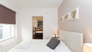 A hotel room with a large bed, a bedside table with a lamp, a view of the bathroom, and a window with blinds.