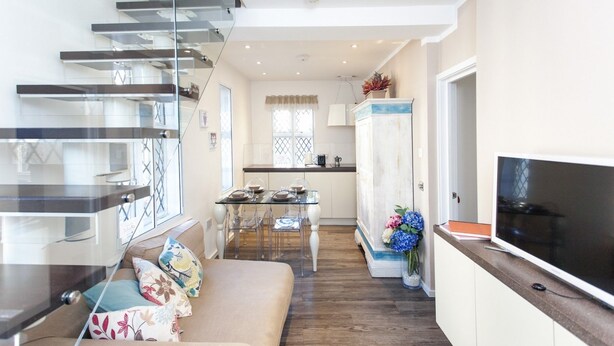 A modern apartment with a glass staircase, a dining area, a kitchen, and a living room with a TV.