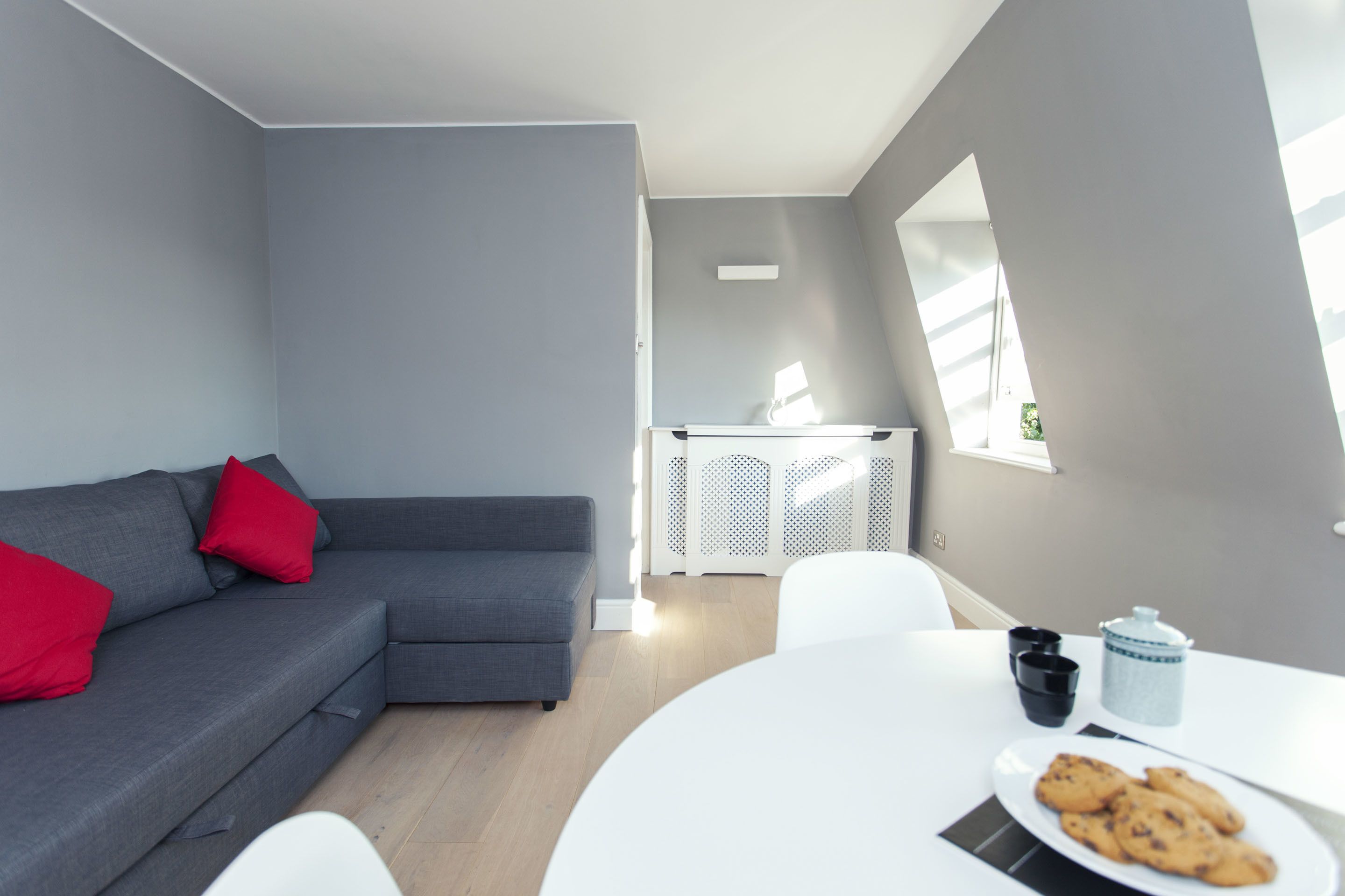 Un salon moderne avec un canapé gris, des coussins rouges, une table blanche sur laquelle se trouve une assiette de biscuits et une fenêtre avec des stores.