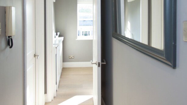 A hallway with a light-colored wooden floor, a large mirror on the right wall, and a door leading to a bathroom. - Shopping Centre and Cinema Atmosphere (London)