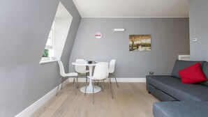 A modern living room with a grey sofa, a dining table with white chairs, a painting on the wall, and a window providing natural light. - Shopping Centre and Cinema Atmosphere (London)