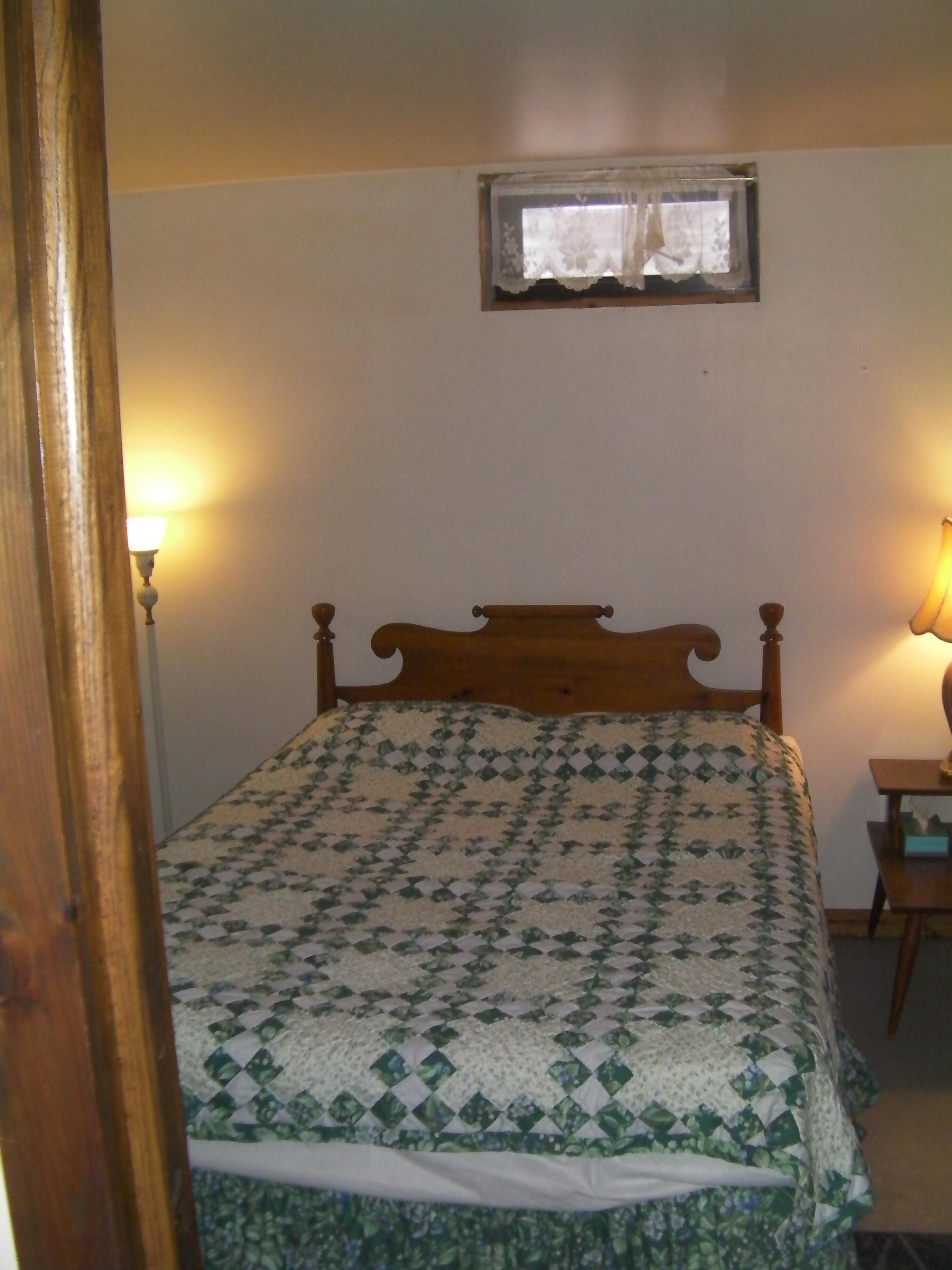 A bedroom with a wooden bed, a quilt, a small window with lace curtains, and two bedside lamps.