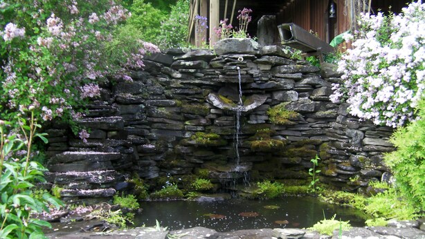 A stone waterfall structure with a pond, surrounded by lush greenery and flowering plants. - Beautiful Getaway - Reynolds Farm Lodge  (Little Marsh)