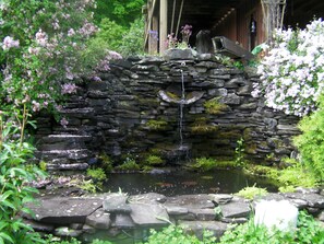 Eine steinerne Wasserfallstruktur mit Teich, umgeben von üppigem Grün und blühenden Pflanzen.