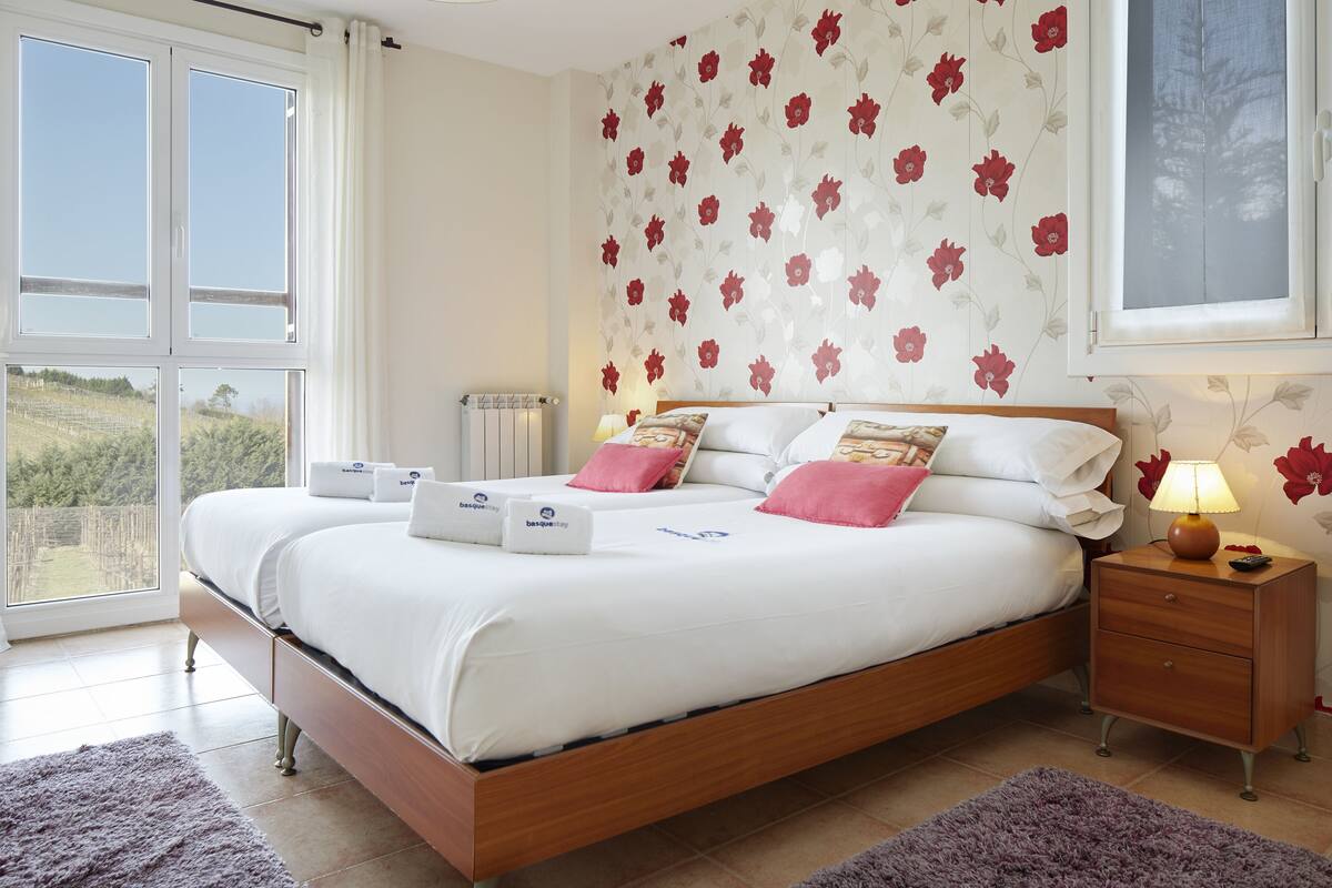 A bedroom with a large bed, a bedside table, a nightstand, a window with curtains, and a floral wallpaper.