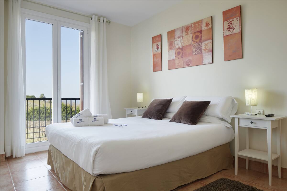 A hotel room with a large bed, two bedside tables, a nightstand, a window with curtains, and artwork on the wall.