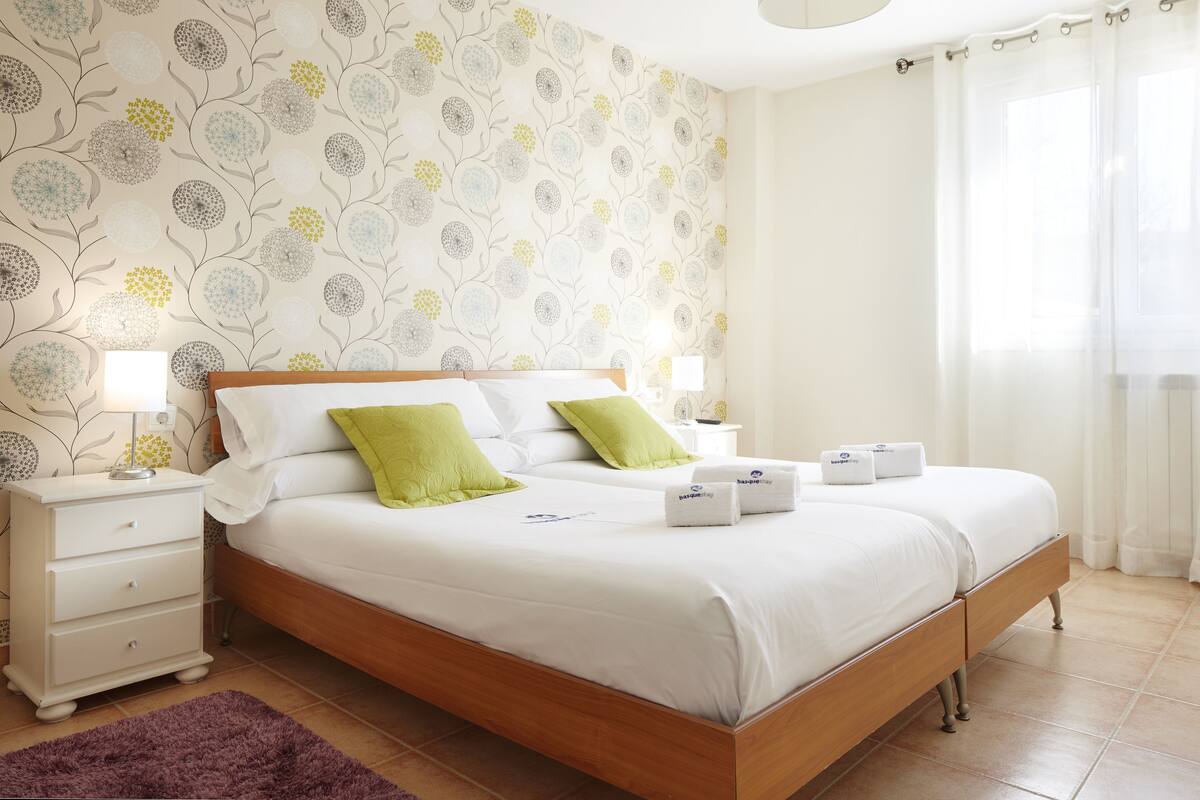 A neatly made double bed with white linens and green pillows, a wooden headboard, a white nightstand with a lamp, and a purple rug on a tiled floor.