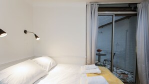 Hotellihuone, jossa on sänky, kaksi yöpöytälamppua, puinen pöytä, jossa on pyyhkeitä, ja näköala parvekkeelle, jossa on pöytä ja tuolit.