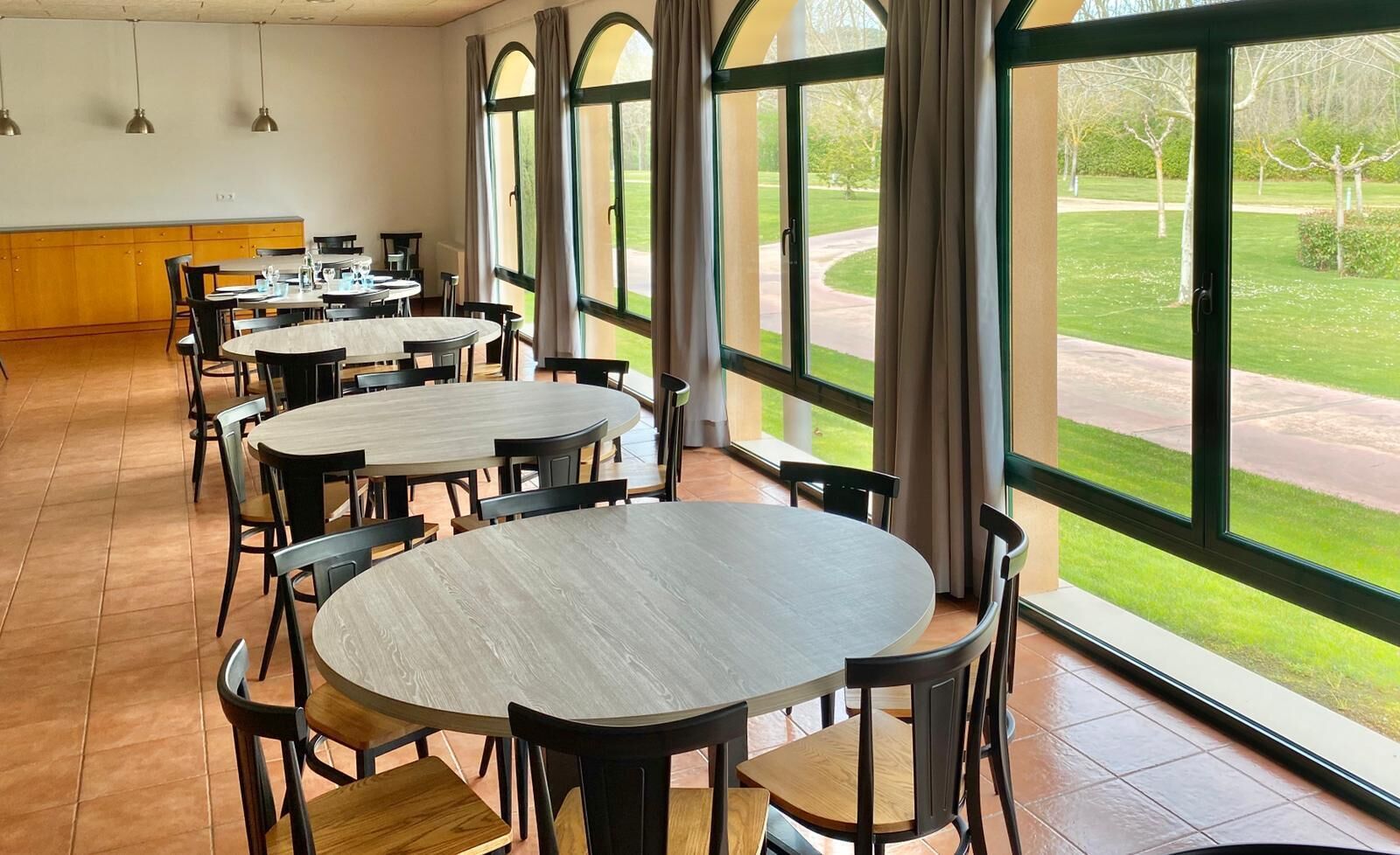 Un comedor amplio con mesas y sillas redondas, ventanas grandes con cortinas, y vistas a un paisaje verde.