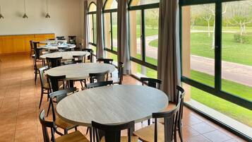 Un comedor amplio con mesas y sillas redondas, ventanas grandes con cortinas, y vistas a un paisaje verde.