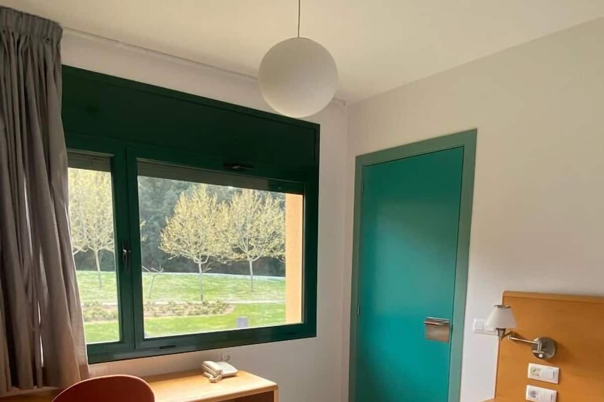 A hotel room with a wooden desk, a chair, a bed, a teal door, and a view of trees through a window.