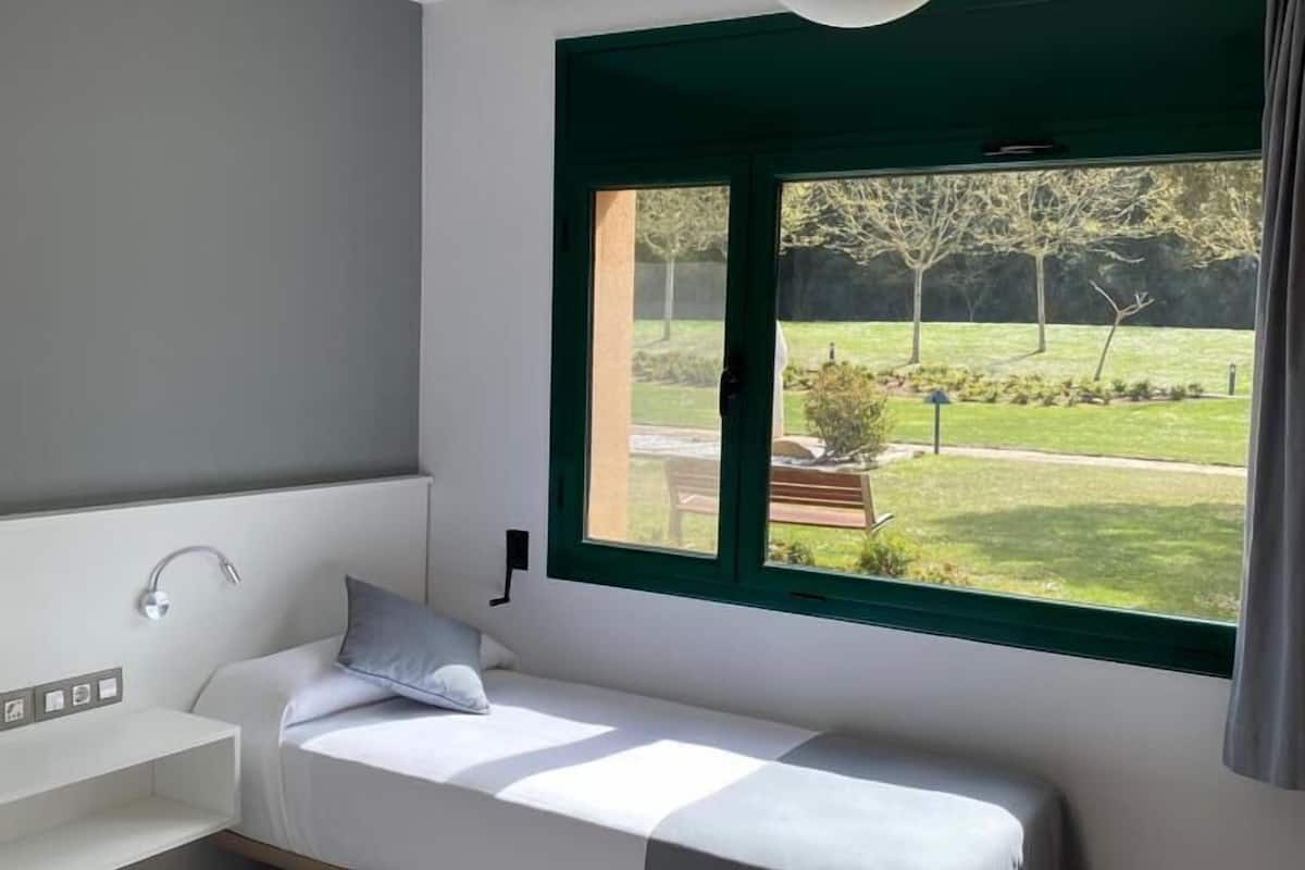 A modern bedroom with a single bed, a small bedside table, a built-in bench, and a large window with a view of a garden.