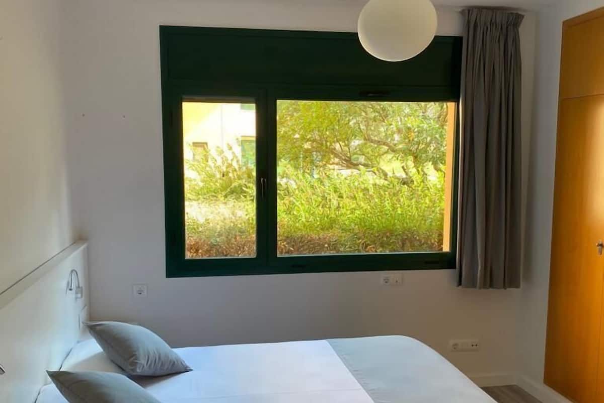 A bedroom with a bed, a nightstand, a window with a view of greenery, and a wooden door.