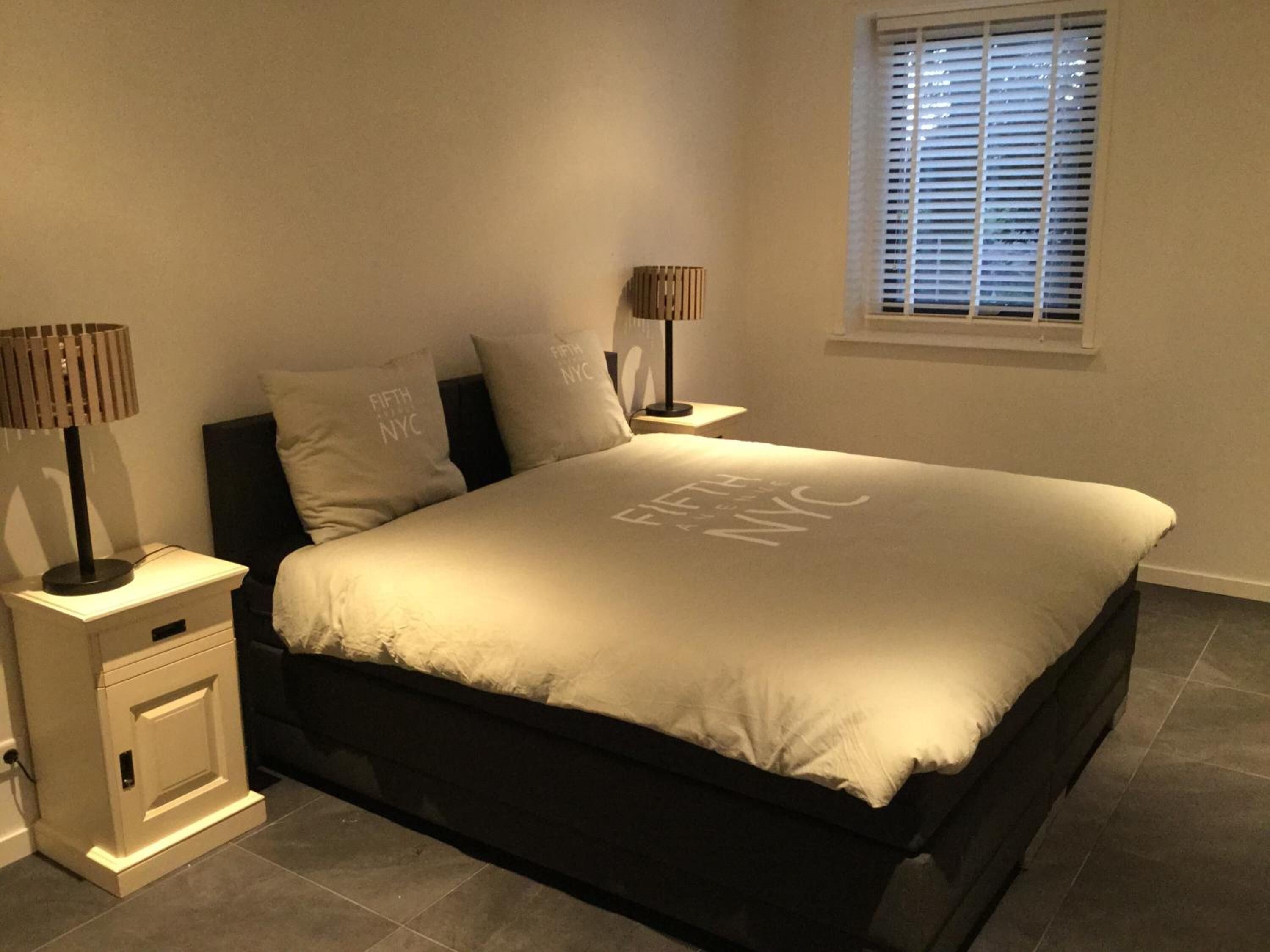 A bedroom with a bed, two bedside tables, a window with blinds, and a nightstand with a lamp.