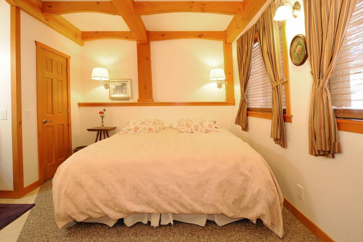 Ein Schlafzimmer mit einem großen Bett, Holzbalken, zwei Wandlampen, einem Fenster mit Vorhängen und einer Tür.