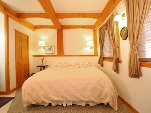 Un dormitorio con una cama grande, vigas de madera, dos lámparas fijadas a la pared, una ventana con cortinas y una puerta.