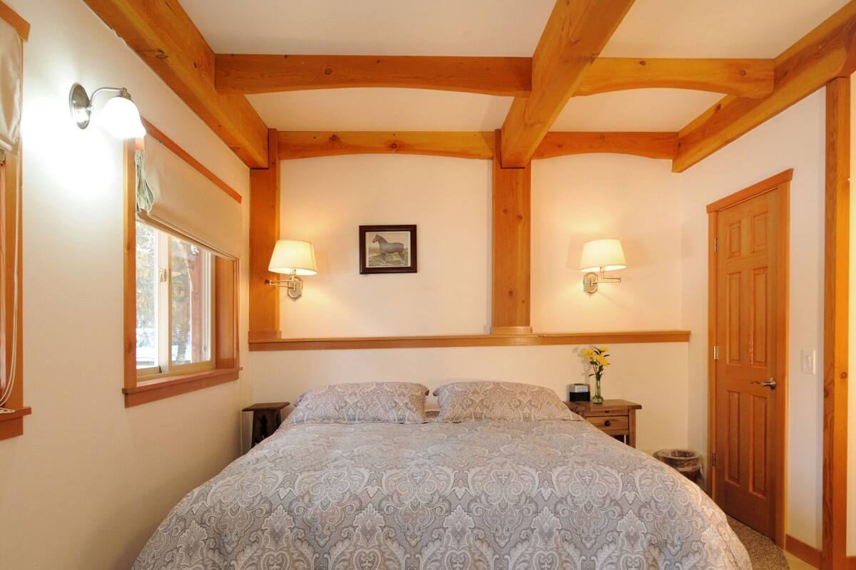 Ein Schlafzimmer mit einem großen Bett, Holzbalken, zwei Wandleuchten, einem gerahmten Bild, einem Nachttisch mit einer Vase und einer Tür.