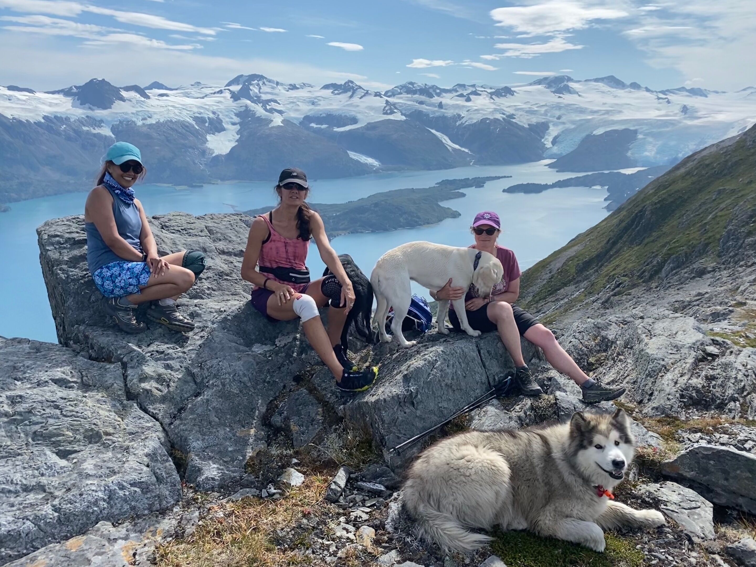 Three people and a dog sitting on a rocky outcrop with a mountainous landscape in the background.