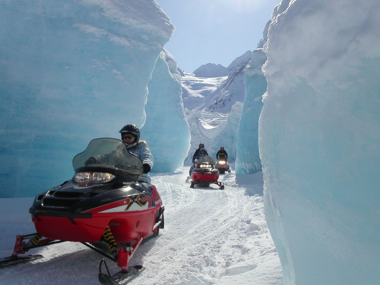 Een smal, met sneeuw bedekt pad, afgegrensd door ijsmuren, met sneeuwscooters en een persoon op een sneeuwscooter.
