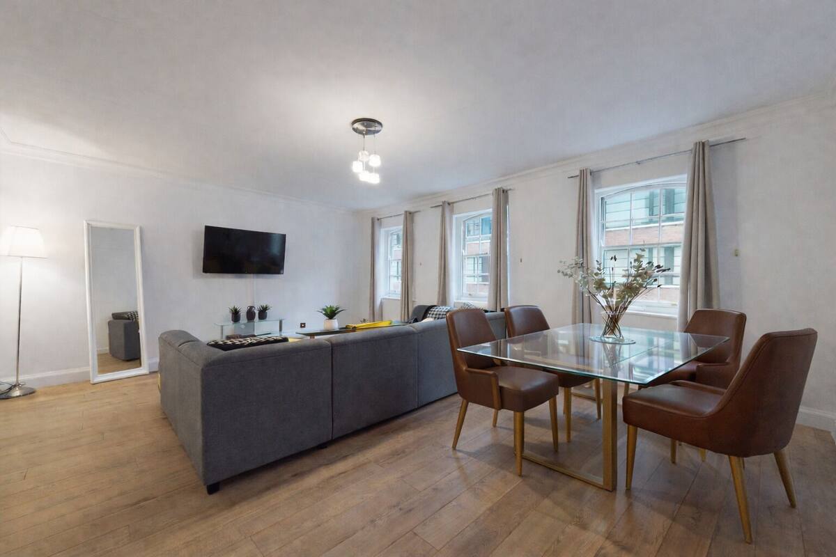 A modern living room with a sofa, a glass dining table with chairs, a flat-screen TV mounted on the wall, and a large window with curtains.