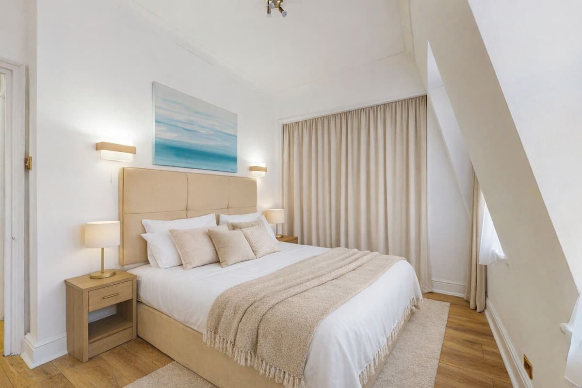 A bedroom with a large bed, two bedside tables, a headboard, a painting, and a window with curtains.