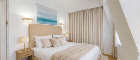 A bedroom with a large bed, two bedside tables, a headboard, a painting, and a window with curtains.