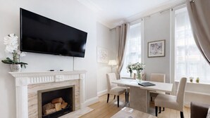 A modern living room with a fireplace, a flat-screen TV, a dining area with chairs, and a coffee table. - Park Lane Apartments - Clarges St (London)