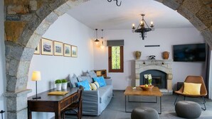 A cozy living room with a stone archway, a fireplace, a sofa, a dining table, and a TV mounted on the wall. - Villa Olga (Agios Nikolaos)